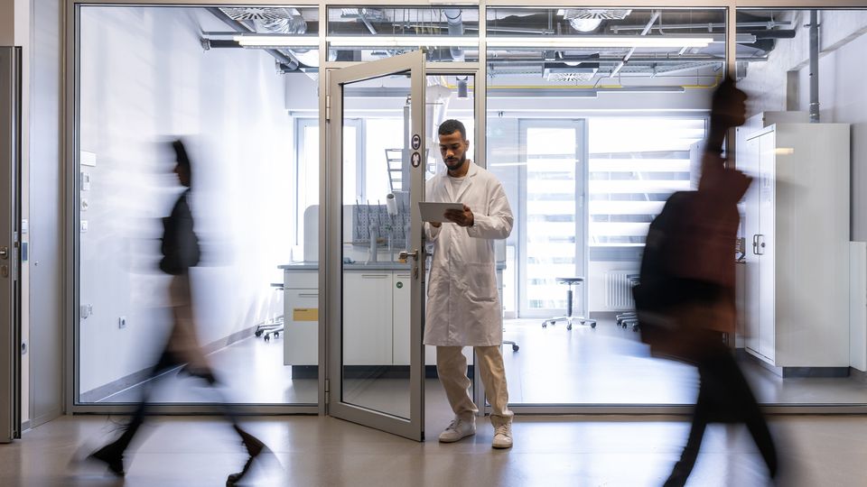 Scientist in a white coat stands between the doors of the faculty laboratory with blurred figures walking past