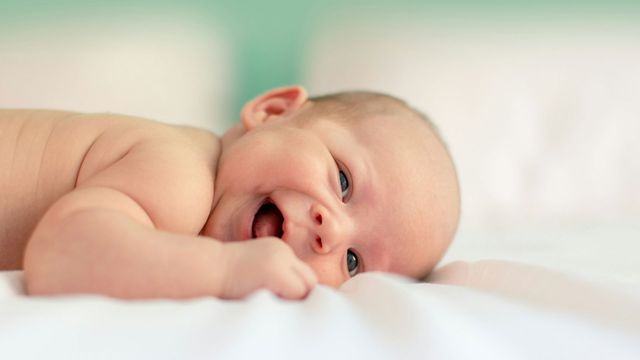 A baby lies on its front, holding a blanket and smiling. 