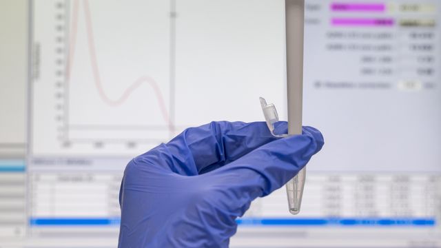Gloved hand holding microcentrifuge tube in front of spectrophotometer data on lab computer screen 