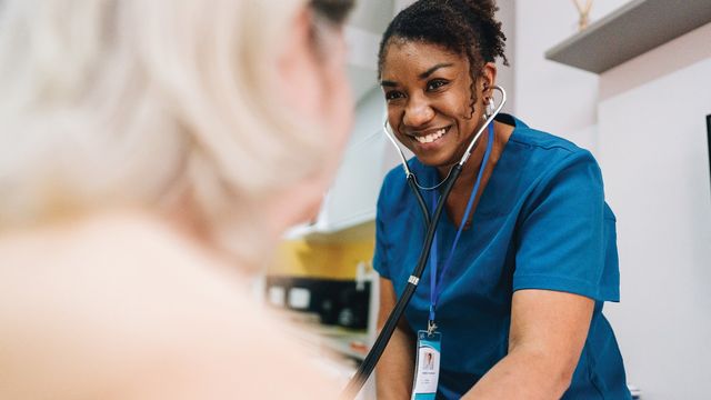 Smiling nurse in scrubs using a stethoscope during a patient consultation, emphasizing compassionate care.